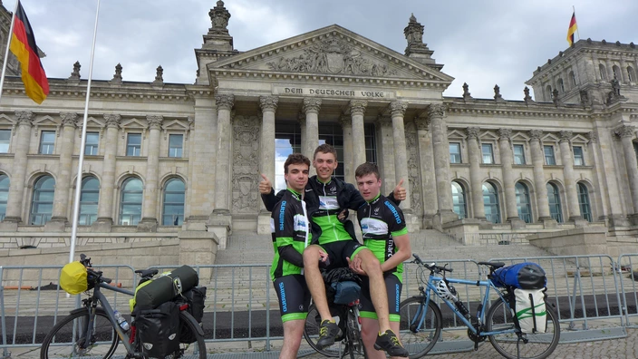 Farbfoto: Drei junge Männer in CJD-Radsportkleidung posieren lachend vor dem Reichstagsgebäude in Berlin. Zwei von ihnen tragen den dritten auf dem Fahrrad sitzend, alle zeigen den Daumen nach oben. Links und rechts stehen voll bepackte Fahrräder mit Gepäcktaschen. Im Hintergrund weht die deutsche Flagge vor dem historischen Gebäude mit der Inschrift „Dem Deutschen Volke“.