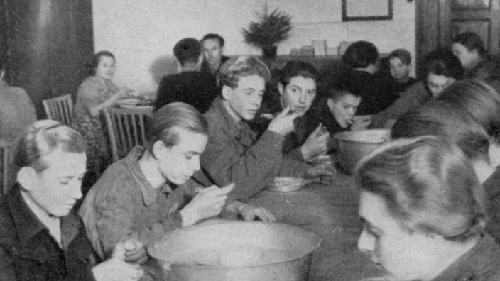 Schwarzweißfoto: Jugendliche sitzen dicht gedrängt an langen Holztischen und essen gemeinsam aus großen Metallschüsseln. Die Szene spielt sich in einem einfachen Speisesaal ab. Im Hintergrund sind weitere Personen an Tischen zu sehen. Die Kleidung und Einrichtung deuten auf die Nachkriegszeit hin, vermutlich 1950er-Jahre. Die Atmosphäre wirkt ruhig und konzentriert.