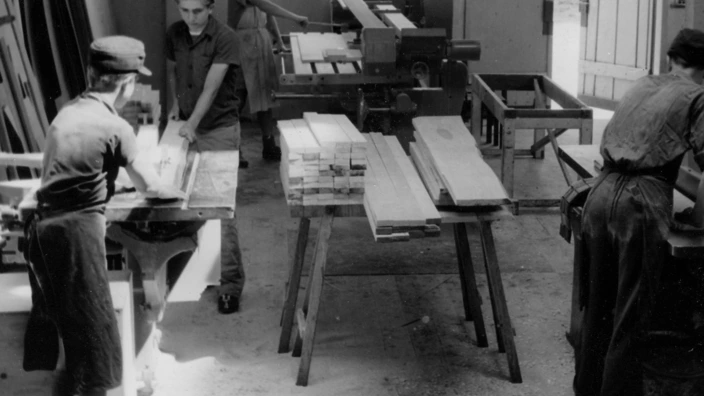 Schwarzweißfoto einer historischen Lehrwerkstatt im Bereich Holzverarbeitung. Mehrere junge Männer arbeiten konzentriert an Werkbänken und Maschinen, sägen und hobeln Holzbretter. Sonnenlicht fällt durch große Fenster auf den Werkstattboden. Die Szene vermittelt eine handwerkliche Ausbildungsatmosphäre der 1950er- oder 1960er-Jahre.