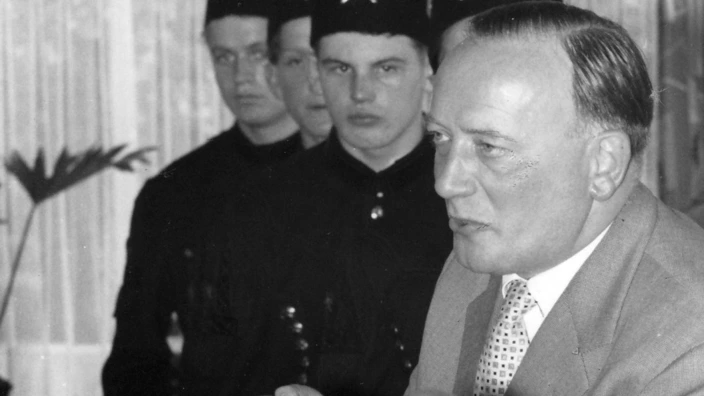 Schwarzweißfoto eines Mannes am Rednerpult mit dem Schild „Jugenddorf“. Hinter ihm stehen vier junge Männer in Bergmannsuniformen mit Federbüschen auf den Hüten. Der Redner – CJD-Gründer Arnold Dannenmann – spricht mit erhobener Hand, im Vordergrund ist ein Mikrofon zu sehen. Die Szene spielt vermutlich bei einer feierlichen Veranstaltung im Rahmen der Berufsausbildung.