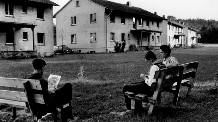 Schwarzweißfoto einer Rasenfläche mit mehreren Bankreihen. Drei Jungen sitzen auf zwei Bänken, lesen oder schreiben. Im Hintergrund stehen einfache Wohnhäuser aus den 1950er-Jahren mit Satteldächern. Links ist ein VW Käfer geparkt. Die Szene wirkt ruhig und alltäglich, im Jugenddorf Dortmund-Oespel.