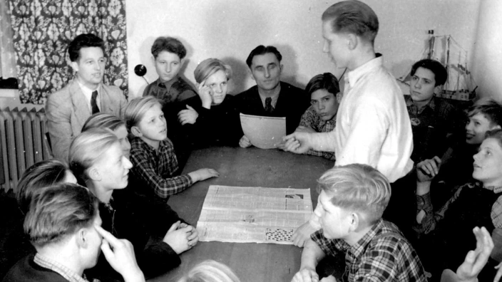 Schwarzweißaufnahme aus den 1950er-Jahren: Etwa 15 Jungen sitzen mit einem Betreuer an einem Tisch in einem schlichten Raum. Einer der Jungen steht und spricht, vermutlich im Rahmen einer Gruppen- oder Mitbestimmungsrunde. Auf dem Tisch liegt eine aufgeschlagene Zeitung. Die Szene vermittelt ein Gefühl von Gemeinschaft, demokratischer Beteiligung und pädagogischer Anleitung.