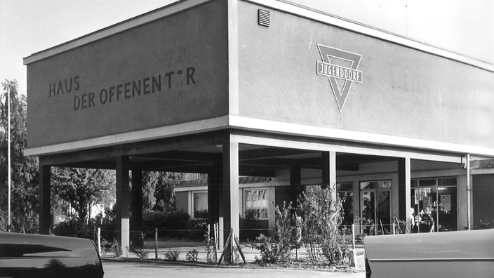 Schwarzweißfoto eines Flachbaus mit moderner Architektur aus den frühen 1960er Jahren. Auf dem Gebäude steht in großen Buchstaben „Haus der offenen Tür“, daneben das markante Dreieckslogo mit der Aufschrift „Jugenddorf“. Vor dem Eingang sind Glasfassade, Vorhänge und vereinzelte Personen zu sehen. Im Vordergrund parken zwei Autos, teilweise angeschnitten. Die Aufnahme entstand in Salzgitter um 1962.