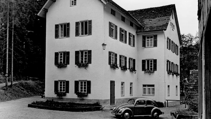 Schwarzweißfoto eines mehrstöckigen, hell verputzten Hauses mit Fensterläden und Blumenkästen, aufgenommen 1954. Das Gebäude ist von dichtem Wald umgeben und steht auf einem gepflasterten Hof. Links im Bild steht ein VW Käfer. Es handelt sich um das Haus Waldfrieden, das erste sozialpädagogische Institut des CJD.