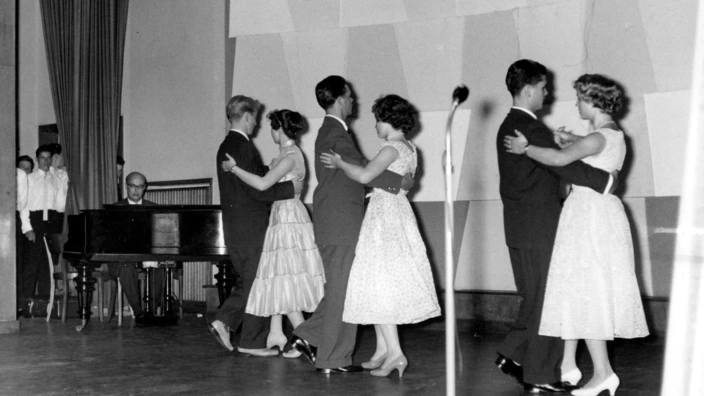 Schwarzweißaufnahme aus dem Jahr 1957: Drei Paare in festlicher Kleidung tanzen Walzer auf einer Bühne während eines musischen Abends im CJD Niederrhein. Im Hintergrund spielt ein Mann Klavier, am Bühnenrand stehen weitere Jugendliche in weißen Hemden.