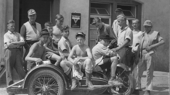 chwarzweißaufnahme aus den frühen Jahren des CJD: Eine Gruppe von etwa fünfzehn Jungen und jungen Männern versammelt sich vor einem Gebäude mit dem Schild "Jugenddorf Neu-Ulrichstein". Einige Jungen sitzen barfuß und teils mit freiem Oberkörper auf einem alten Motorrad mit Seitenwagen, andere stehen im Hintergrund. Die Szene wirkt sommerlich und entspannt.