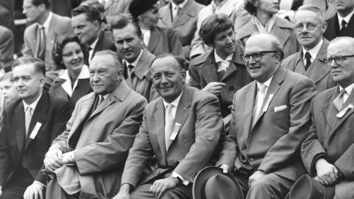Schwarz-Weiß-Foto von Prominenten in einer Zuschauertribüne, Bonn 1957: In der ersten Reihe sitzen (von links nach rechts) mehrere Männer in Anzügen, darunter Bundeskanzler Konrad Adenauer (zweiter von links) und Arnold Dannenmann (Mitte), alle mit Ehrenbändchen am Revers. Die Männer tragen Anzug und Mantel, einige halten ihre Hüte auf dem Schoß. Im Hintergrund weitere Zuschauerinnen und Zuschauer. Das Bild entstand beim Sportfest 1957 in Bonn.
