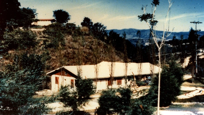 Farbfoto eines Hauses in hügeliger Landschaft: Ein einfaches Gebäude mit Satteldach steht eingebettet in eine bewaldete Berglandschaft. Es handelt sich um ein Haus des CJD in Los Teques, Venezuela, das zwischen 1951 und 1960 betrieben wurde. Im Vordergrund junge Bäume, im Hintergrund bewaldete Hänge und eine weite Sicht auf entfernte Gebirgszüge.
