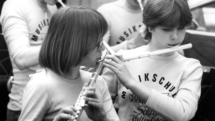 Schwarz-weiß-Fotografie einer Gruppe junger Menschen, die Querflöte spielen. Auf ihren Pullovern steht „Musikschule Jugenddorf Schloss Kaltenstein“. Zwei Mädchen im Vordergrund sind konzentriert auf ihr Spiel.