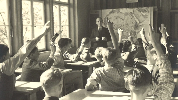 Schwarz-weiß-Fotografie eines Klassenzimmers aus der Mitte des 20. Jahrhunderts: Schüler mit erhobenen Händen sitzen an Holzbänken, während ein Lehrer vor einer Weltkarte unterrichtet.