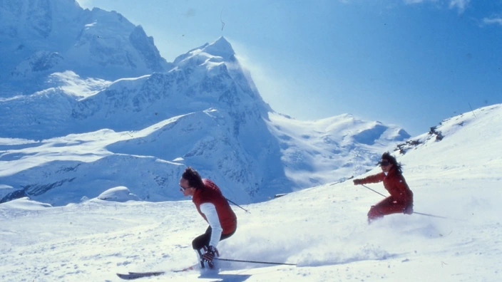 Zwei Personen in roten Skianzügen fahren bei strahlendem Sonnenschein eine verschneite Piste hinab. Im Hintergrund erheben sich majestätisch schneebedeckte Berggipfel.