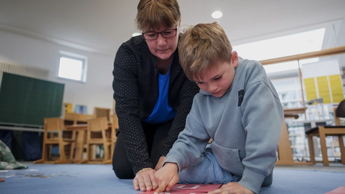 Eine Frau kniet neben einem Jungen auf dem Boden eines Klassenraums. Gemeinsam zeigen sie auf eine Lernkarte mit dem Buchstaben „N“. Im Hintergrund stehen Stühle und Tische.