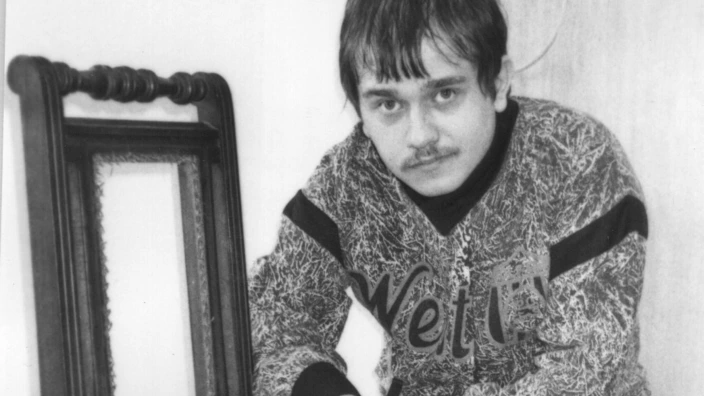 Schwarz-weiß-Fotografie eines jungen Mannes, der auf einem Stuhl sitzt und den Sitz einer antiken Holzstuhllehne mit Geflecht erneuert. Er trägt einen gemusterten Pullover und schaut direkt in die Kamera.