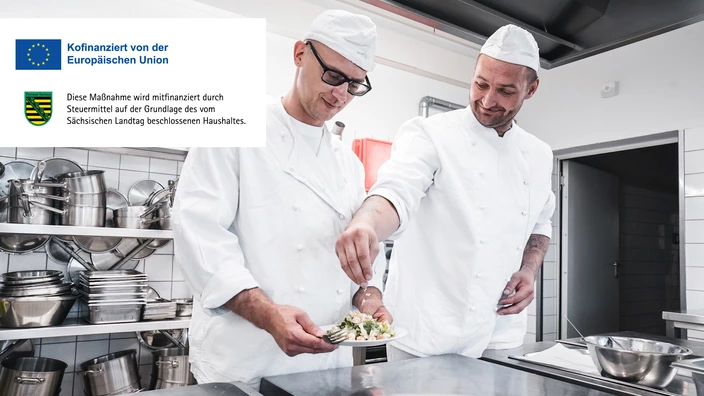 Zwei Männer mit Kochkleidung garnieren eine Mahlzeit auf einem Teller. 