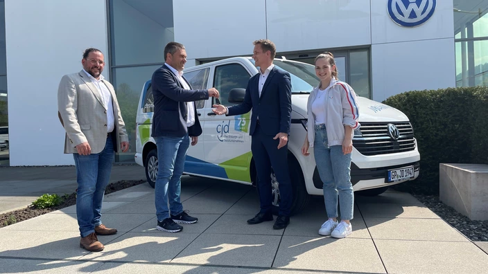 Vier Personen stehen vor einem weißen VW-Transporter mit CJD-Branding und dem Logo „75 Jahre CJD – Das Bildungs- und Sozialunternehmen“. Zwei Männer in der Mitte reichen sich symbolisch den Autoschlüssel zur Übergabe. Im Hintergrund das Gebäude eines Volkswagen-Autohauses mit großem VW-Logo. Die Szene findet bei sonnigem Wetter statt.