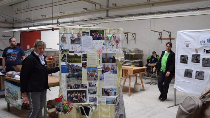 In einer Werkhalle präsentieren mehrere Personen kreative Arbeiten und Projektinformationen. Im Vordergrund steht eine Frau neben einer großen Stellwand, die mit Fotos, Texten und handgefertigten Dekorationen bestückt ist. Rechts daneben ist eine weitere Infotafel mit dem CJD-Logo zu sehen. Im Hintergrund befinden sich Werkbänke, Regale und weitere Personen, die die Ausstellung betrachten oder betreuen.