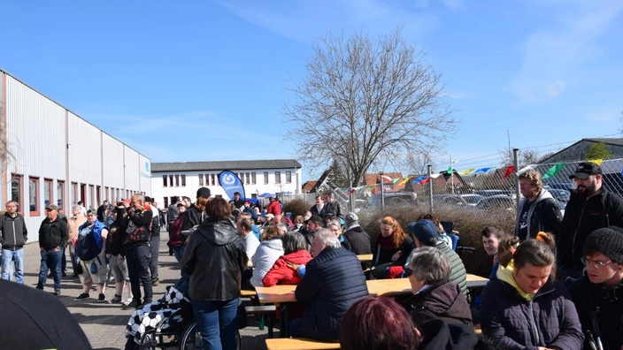 Bei sonnigem Wetter sitzen zahlreiche Menschen an langen Holzbänken im Freien. Einige unterhalten sich, andere blicken in Richtung Kamera. Im Hintergrund stehen weitere Personen, teils vor einer großen Halle mit roten Fenstern. Bunte Wimpelketten schmücken den Zaun, dahinter parken Autos. Die Atmosphäre wirkt gesellig und lebendig, als Teil einer Veranstaltung.