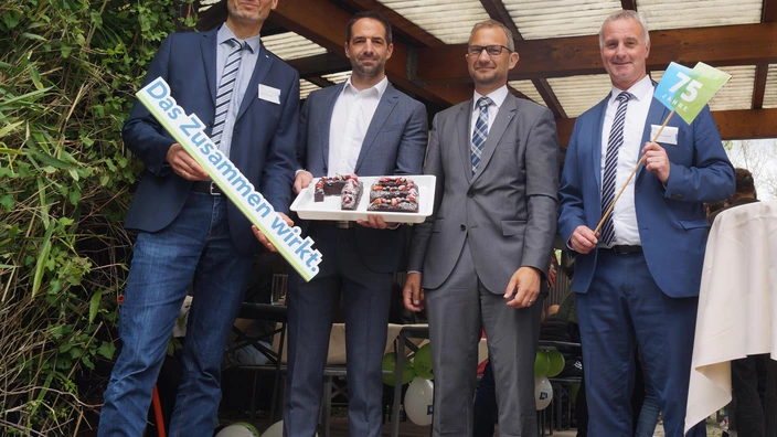Vier Männer in Anzügen stehen nebeneinander und lächeln in die Kamera. Der Mann links hält ein Schild mit der Aufschrift „Das Zusammen wirkt.“, der zweite von links präsentiert ein Tablett mit Kuchenstücken in Form der Zahl „75“, verziert mit Beeren. Der Mann rechts hält ein kleines Fähnchen mit „75 Jahre“. Im Hintergrund sind Luftballons und weitere Gäste unter einer überdachten Terrasse zu sehen.