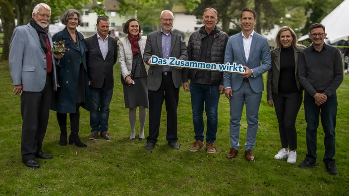 Neun Personen stehen in einer Reihe auf einer Wiese im Freien, einige von ihnen lächeln in die Kamera. Zwei Männer in der Mitte halten ein Schild mit der Aufschrift „Das Zusammen wirkt.“. Die Gruppe ist warm gekleidet, im Hintergrund sind Bäume, Rasenflächen und ein weißes Zelt zu sehen.