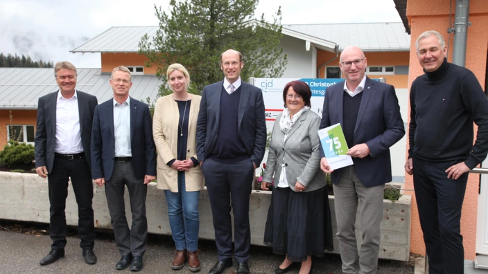 Sieben Personen stehen in einer Reihe vor einem Gebäude des CJD, erkennbar am Schild „Christophorusschule“. In der Mitte hält ein Mann ein Schild mit der Aufschrift „75 Jahre“. Die Gruppe besteht aus Männern und Frauen in formeller Kleidung, die lächelnd in die Kamera blicken. Im Hintergrund sind orangefarbene Gebäudeteile, ein Baum sowie Berge im Nebel zu sehen.