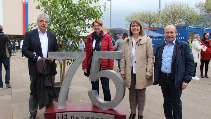 Vier Personen stehen im Freien neben einer großen silbernen Skulptur der Zahl „75“ mit der Aufschrift „cjd – Das Zusammen wirkt.“ auf dem Sockel. Die Gruppe lächelt in die Kamera, im Hintergrund sind weitere Menschen, ein Baum, Container und Gebäude zu sehen. Die Szene wirkt wie eine Jubiläumsfeier oder ein öffentlicher Anlass.
