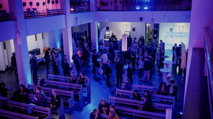 Blick von einer Empore auf eine große Halle mit Kirchenbänken und vielen Menschen, die in lockeren Gruppen stehen, reden und Getränke in der Hand halten. Im Hintergrund sind eine Bar und Stehtische aufgebaut. Der Raum ist in blau-violettes Licht getaucht und vermittelt eine lebendige, festliche Atmosphäre.