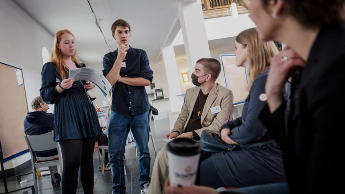 Eine Gruppe junger Menschen sitzt in einem Stuhlkreis in einem hellen Raum und diskutiert. Zwei Personen stehen, eine Frau hält ein Blatt Papier, ein Mann neben ihr spricht nachdenklich. Die anderen Teilnehmenden hören aufmerksam zu, eine Person trägt eine Maske und ein Kreuz um den Hals. Im Vordergrund ist ein Kaffeebecher zu sehen. Im Hintergrund hängen leere Pinnwände für die weitere Arbeit.