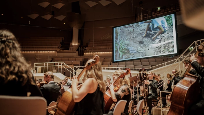Ein Orchester spielt in einem Konzertsaal, im Vordergrund sind Musikerinnen mit Celli und Kontrabass zu sehen. Im Hintergrund hängt eine große Leinwand, auf der ein Film mit einer Person bei der Arbeit im Erdreich gezeigt wird. Die Musiker tragen Konzertkleidung, die Atmosphäre wirkt konzentriert und festlich.