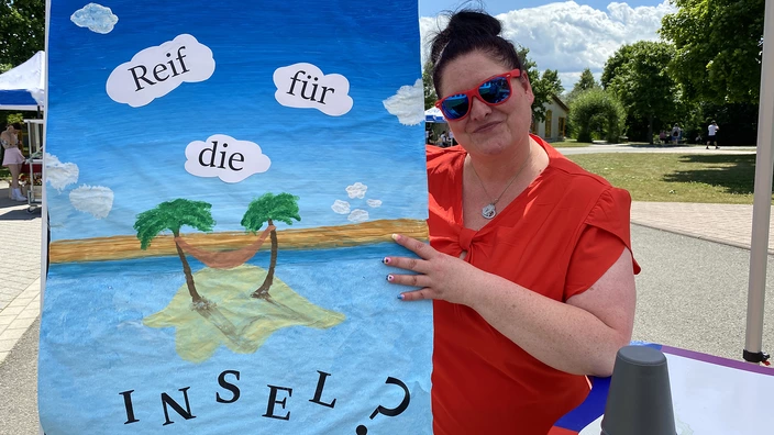 Lydia Motz steht neben einem bunten, selbstgemalten Plakat mit der Aufschrift "Reif für die Insel?"