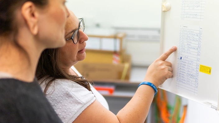 Zwei Personen stehen vor einer weißen Schranktür, an der ein Plan befestigt ist. Eine Person zeigt mit der rechten Hand auf eine Liste auf dem Plan. Der Inhalt ist nicht lesbar. Im Hintergrund sind einige Büroartikel und Kartons zu erkennen.