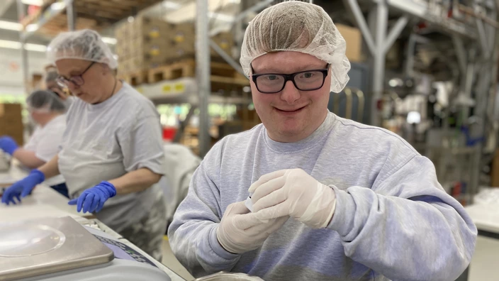 Zwei Personen arbeiten in einem Produktions- oder Verpackungsbereich. Beide tragen Haarnetze, Handschuhe und Schutzkleidung. Im Vordergrund sortiert eine Person kleine weiße Gegenstände aus einer großen Schale. Im Hintergrund sind Regale mit Kartons und industrielle Geräte zu sehen. Die Arbeitsumgebung wirkt sauber und organisiert.