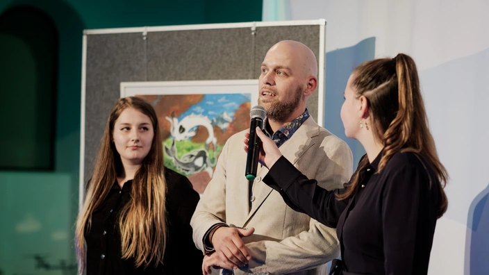  Stephan Zwingmann spricht bei der Ausstellung im Rahmen des CJD Orchesterkonzerts 2023 im Foyer der Berliner Philharmonie in ein Mikrofon, das von einer jungen Frau rechts neben ihm gehalten wird. Links neben ihm steht eine weitere junge Frau. Im Hintergrund hängt ein Bild aus der Ausstellung.