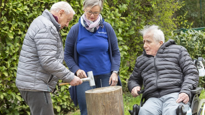 2 Herren mit Jacke und eine Dame mit Strickjacke versuchen im Garten 3 Nägel mit einem speziellen Hammer in einem Holzblock zu versenken.