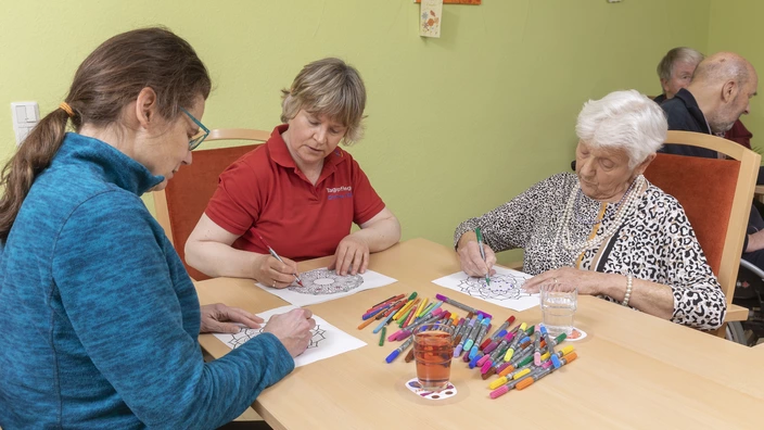 3 Frauen sitzen an einem Tischen und malen Mandalas mit vielen bunten Filzstiften Mandalas aus.