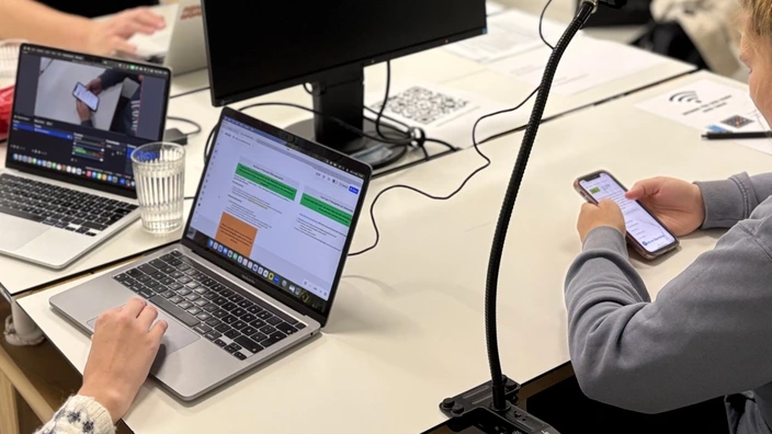 Eine Person bedient ein Smartphone und daneben sind zwei offene Notebooks zu sehen an denen eine weitere Person arbeitet.