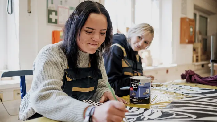 Produktionsschule Wolgast: Junge Menschen arbeiten gemeinsam in einer hellen Werkstatt. Im Vordergrund sitzt eine junge Frau, konzentriert bei der Arbeit, während im Hintergrund eine Betreuerin das handwerkliche Geschehen begleiten. Das Bild vermittelt Teamgeist, handwerkliches Lernen und die unterstützende Atmosphäre in einer CJD Produktionsschule.