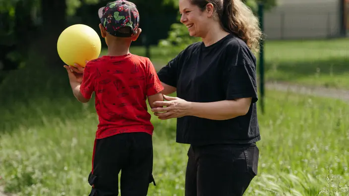 Ein Kind balaciert mit einem gelben Ball über eine Turnbank. Eine Pädagogin läuft nebenher und berührt das Kind am Arm.