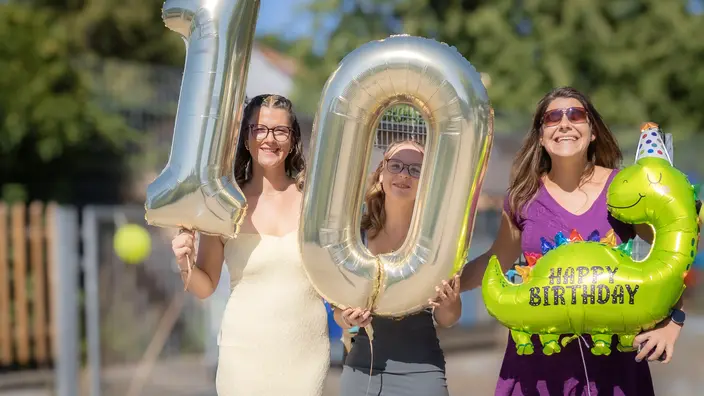 3 Personen halten große Heliumballons in den Händen. Zu sehen ist die Zahl 10 und ein grüner Dinosaurier mit der Aufschrift "Happy Birthday".