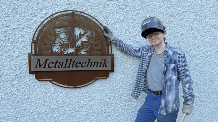 Maciej Lehr steht mit hochgeklappter Schweißmaske und Werkzeug in der linken Hand vor einer weißen Hauswand rechts neben einem runden Schild aus Metall auf dem Metalltechnik steht.