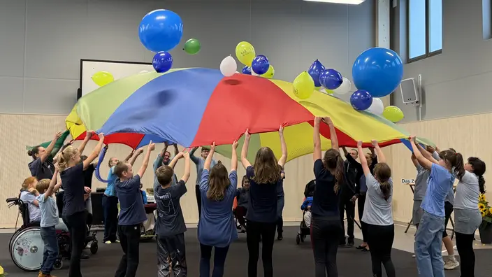 Schüler und Pädagogen werfen mit einem großen, bunten Schwungtuch Luftballons nach oben.