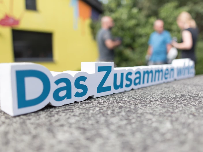 Schriftzug „Das Zusammen wirkt.“ im Vordergrund, unscharfe Personen im Hintergrund, Gemeinschaft beim CJD.