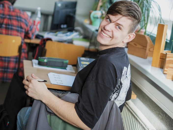 Ein junger Mann sitzt in einem Klassenraum oder Hörsaal, bearbeitet ein Arbeitsblatt und lacht in die Kamera.