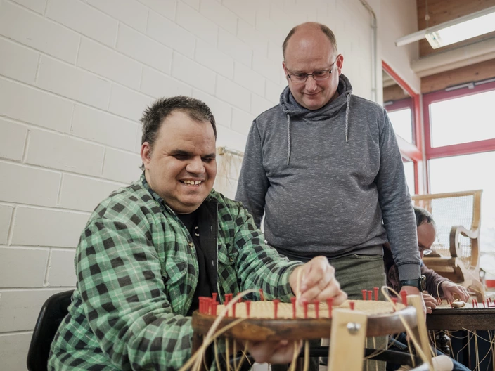 Ein Mann mit Behinderung restauriert den Sitz eines Stuhles in einer Werkstatt. Ein anderer Mann sieht ihm dabei zu. Spenden helfen Menschen mit Behinderung die Teilhabe am Arbeitsmarkt zu ermöglichen.