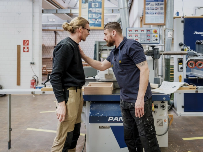 Ein Mann ermutigt einen jüngeren Mann und legt ihm seinen Arm auf die Schulter. Sie arbeiten in einer Holz-Werkstatt des CJD.
