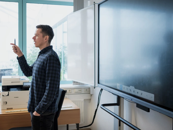 Ein Lehrer steht vor einer digitalen Tafel, hält einen Stift hoch und spricht.