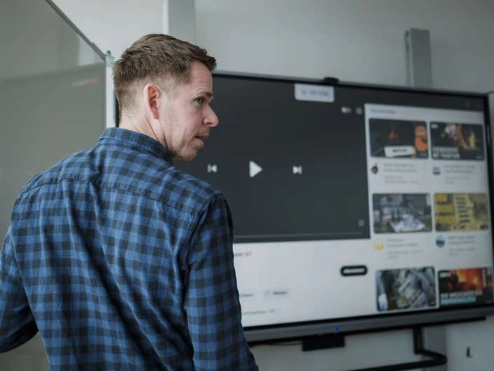 Junger Lehrer steht mit dem Rücken zur Klasse an einer digitalen Tafel und zeigt ein Video.