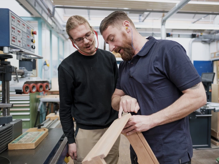 In einer Werkstatt erklärt ein Ausbilder einem Auszubildenden etwas an einem Holzrahmen, den er in der Hand hält. Durch Spenden kann jungen Menschen geholfen werden ihren beruflichen Weg zu finden.