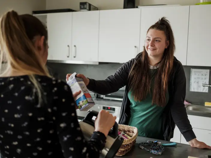 Eine junge Frau reicht in einer Küche eine Packung Milch an eine andere junge Frau, die ihr gegenübersteht und gerade mit einer Kanne etwas eingießt. Spenden unterstützen junge Menschen in Wohngruppen des CJD.