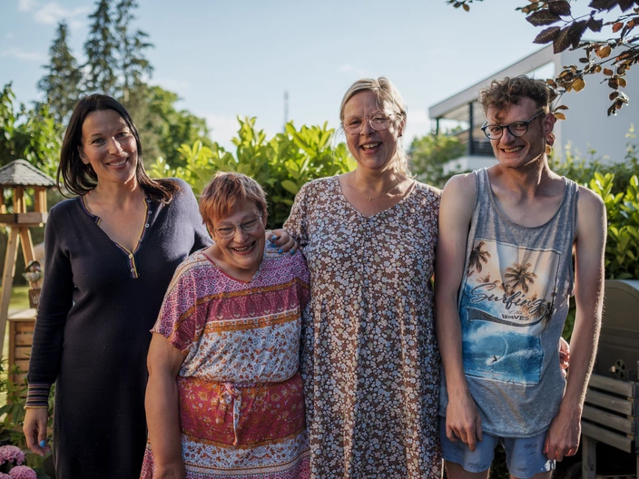 Vier Menschen – zwei Frauen, eine jüngere Frau und ein junger Mann – stehen entspannt im Garten, lächeln herzlich in die Kamera und genießen die sommerliche Atmosphäre. Bei uns stehen Inklusion, Zusammenhalt und Lebensfreude im Mittelpunkt. Erlebe unser vielfältiges Miteinander – jetzt über unsere inklusiven Angebote informieren und mitmachen!