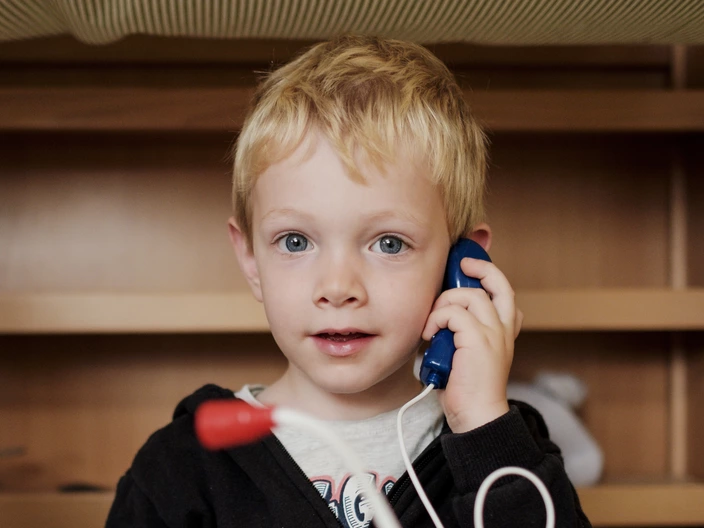 Ein blondhaariger Junge hält ein blaues Spielzeugtelefon ans Ohr und schaut aufmerksam in die Kamera. Vor ihm liegen weitere Kabel und Spielzeuge auf dem Tisch.